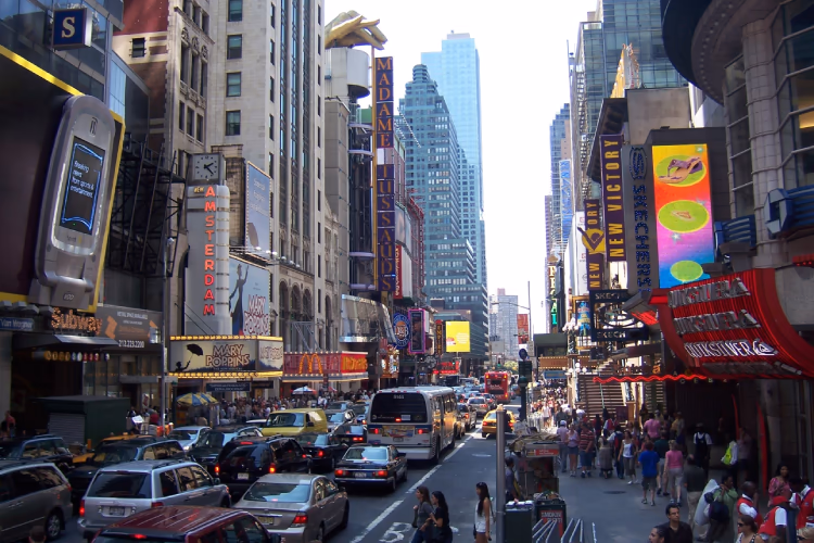 A street with two lanes of mixed-traffic in either direction, and third bus/taxi lane. Pedestrians are squashed into the fairly narrow pavements on each side, with a few waiting to cross the backed-up lanes of traffic. The cars undoubtedly dominate this street, despite the many many shops, restaurants and attractions on it, and even a subway station on the left side.
