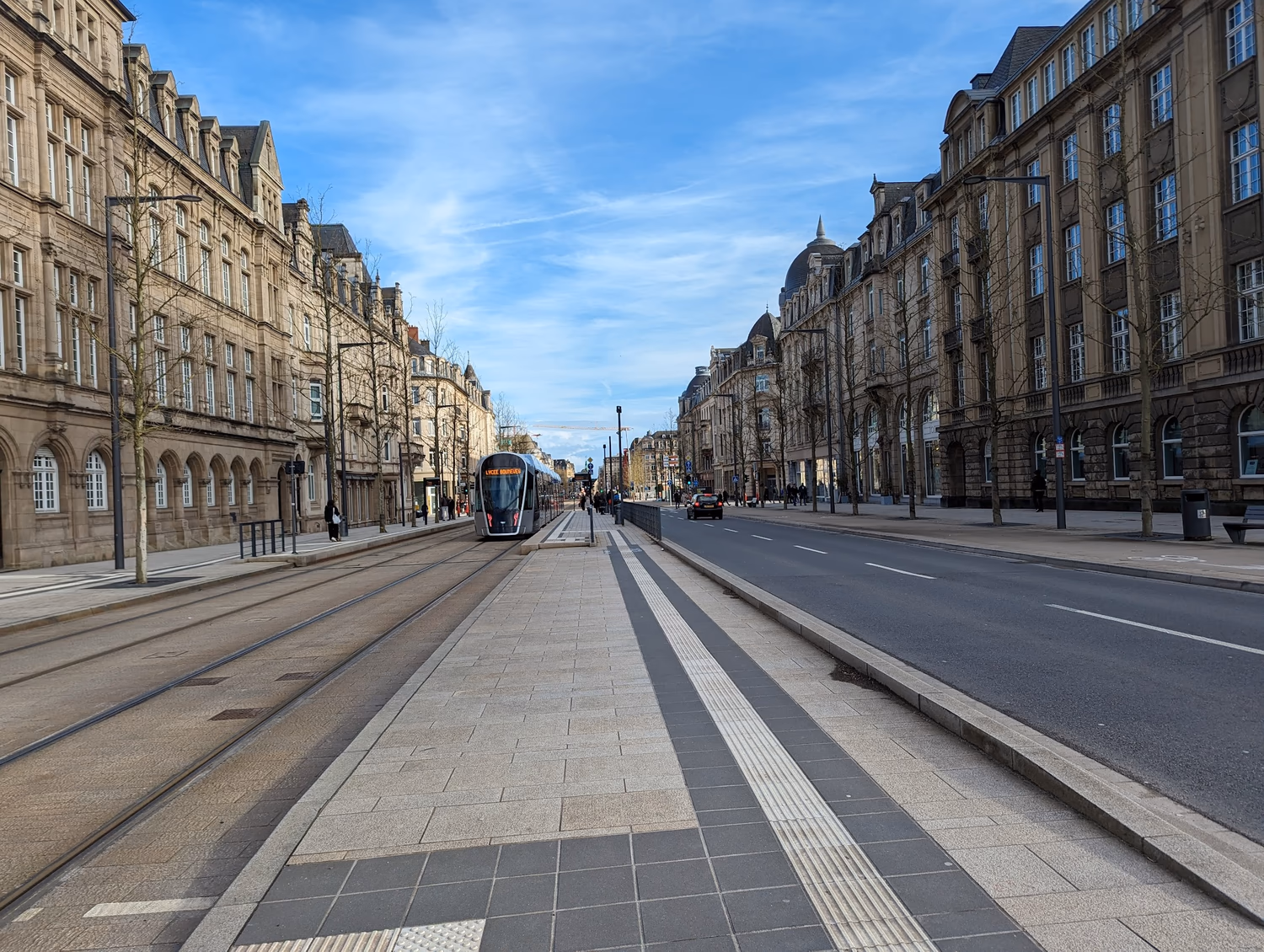 A photo I took standing in the middle of one of the main streets in Luxembourg. On either side are shops of about 4 floors, which are a maybe 2/5 the height of the width of the street. There are quite wide pavements, and on the left two trams lanes and a tram in shot, and more pavement and tramstops in the centre. To the right are two reasonably narrow road lanes and to its right a cycle path. The buildings and street (although not the road) are yellow-grey and well-kept, with trees, benches, and bins.